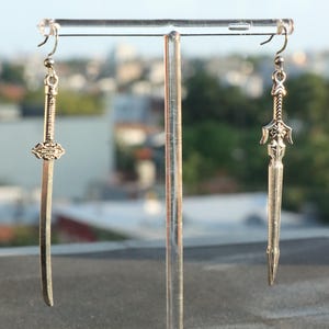 May include: A pair of silver-toned metal earrings shaped like swords. One earring features a long, slender blade with a detailed hilt, while the other earring displays a broader, ornate blade design with a crossguard. Both hang from simple silver hooks.
