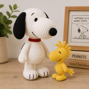 May include: A white and black crocheted dog figurine with a red collar standing next to a smaller yellow crocheted bird figurine. Both figures are crafted with visible yarn stitches and have embroidered facial features, standing on a wooden surface.