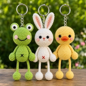 May include: Three crocheted keychains featuring a green frog, a white rabbit, and a yellow duck. Each figure has black bead eyes, pink stitched cheeks, and a small embroidered cross on the belly. The figures are attached to silver metal key rings.