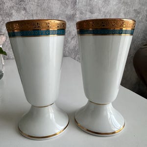 Può includere: Due calici in porcellana bianca con bordo svasato e base a piedistallo. Il bordo superiore è decorato con una fascia di intricati motivi floreali dorati e una striscia color ottanio. La base di ogni calice è rifinita con un sottile filo dorato.