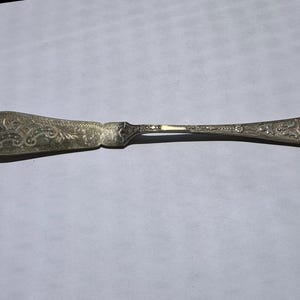 Puede incluir: Un cuchillo para mantequilla de metal plateado con un patrón grabado y ornamentado en la hoja ancha y plana y en el mango delgado. El diseño incluye intrincados arabescos y motivos decorativos, con una zona lisa en forma de escudo.