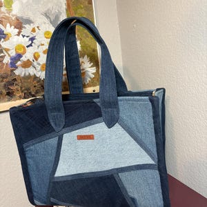 May include: A tote bag constructed from patchwork denim fabric in varying shades of dark and light blue. The bag features two sturdy denim handles, a rectangular silhouette, and a small brown rectangular label centered on the front panel.