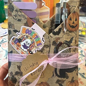 May include: A paper envelope with a vintage newspaper and pumpkin print, tied with a pink ribbon and a tag labeled Witchy Romance. It contains stickers of a ghost in a bookstore and a coffee cup, plus pens and a wax seal stamp.