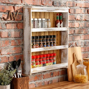 May include: A wall-mounted wooden spice rack with a distressed white finish and natural wood back panel. It features three shelves holding various glass spice jars and two hot sauce bottles. The rack is positioned against a red brick wall.