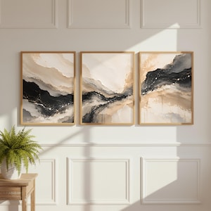 May include: A set of three vertical wall art prints in light wood frames, featuring abstract fluid designs in black, beige, cream, and white with subtle gold-toned accents, displayed side-by-side on a wall with decorative molding.