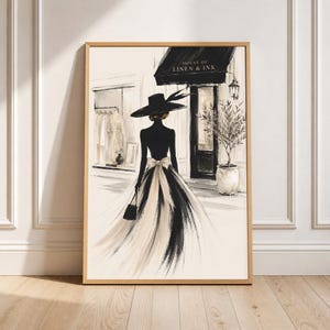 Könnte beinhalten: Ein gerahmter Kunstdruck mit einer Modeillustration einer Figur mit schwarzem Hut mit breiter Krempe und einem fließenden schwarz-weißen Rock mit Schleife, die vor einem Geschäft namens HOUSE OF LINEN & INK mit einem Olivenbaum im Topf steht.