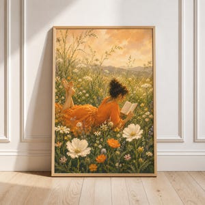 May include: A rectangular wall art print in a light wood frame depicting a person with dark hair in an orange dress lying in a field of white and orange wildflowers, reading an open book under a warm, golden-toned sunset sky with distant hills.