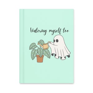 Könnte beinhalten: Ein helles mintgrünes Notizbuch mit festem Einband und einer Cartoon-Illustration eines weißen Geistes, der eine beige Gießkanne hält und eine grüne Topfpflanze gießt. Der Text "Watering myself too" ist in schwarzer Schrift abgebildet.