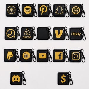 May include: A collection of square black keychains featuring gold-colored icons representing various social media and web platforms, including Wi-Fi, Spotify, Pinterest, Snapchat, Globe, Airbnb, Vimeo, eBay, PayPal, LinkedIn, YouTube, Facebook, Instagram, Discord, and a dollar sign.
