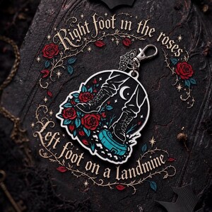 May include: A circular metal keychain featuring a graphic of two boots, one stepping on red roses and the other on a blue landmine. The text reads Right foot in the roses and Left foot on a landmine. It includes a silver-toned lobster clasp attachment.