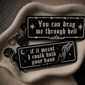 May include: Two rectangular black keychains with white text and graphics. One reads 'You can drag me through hell' with star accents. The other reads 'if it meant I could hold your hand' with a skeleton hand reaching for a human hand and moon icons.