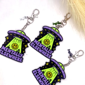 May include: Three keychains featuring a purple and black UFO beaming down a yellow smiley face. The text MENTALLY ELSEWHERE is printed in purple. Each keychain includes a silver metal lobster clasp and a small alien charm in white, green, or blue.
