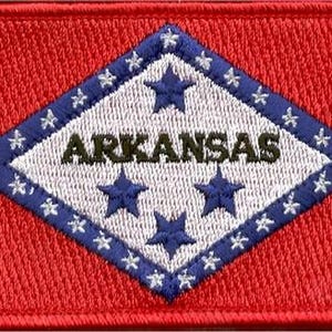 May include: A rectangular embroidered patch featuring the Arkansas state flag design. The background is bright red with a central white diamond bordered in blue. The word ARKANSAS is stitched in dark blue, surrounded by blue stars.