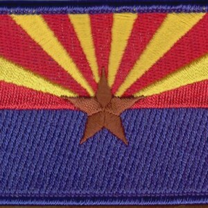 May include: A rectangular embroidered patch featuring the Arizona state flag design. The upper half displays thirteen alternating red and yellow rays, while the lower half is solid blue. A large, five-pointed copper-colored star is centered.