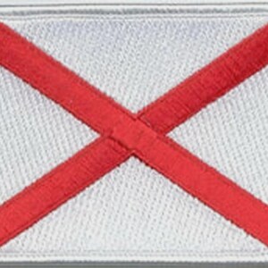 May include: A rectangular embroidered patch featuring a white background with a red saltire cross extending from corner to corner. The patch has a white stitched border around the entire perimeter, creating a clean and defined edge for the design.