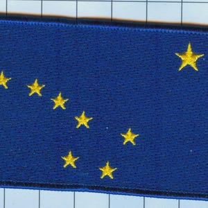 Puede incluir: Un parche rectangular bordado que presenta un fondo azul profundo con ocho estrellas amarillas dispuestas en el patrón de la Osa Mayor y la Estrella Polar, representando la bandera de Alaska, con un borde cosido de color azul oscuro.