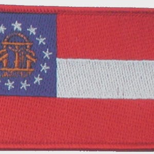 Puede incluir: Un parche bordado rectangular que muestra la bandera del estado de Georgia. El diseño incluye una franja vertical azul a la izquierda con un sello estatal dorado rodeado por trece estrellas blancas, y tres franjas horizontales roja, blanca y roja.