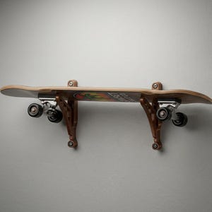 Puede incluir: Un monopatín montado horizontalmente en una pared mediante dos soportes de madera de color marrón oscuro. El monopatín presenta una tabla de madera natural con ruedas negras y ejes de metal plateado. Se ve un gráfico de palmera.