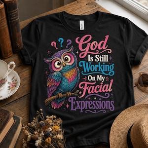 May include: A black t-shirt featuring a colorful owl illustration perched on a branch with the text "God Is Still Working On My Facial Expressions" in pink and blue script and block lettering, accompanied by three small question marks above.