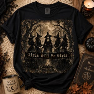 May include: A black short-sleeved t-shirt featuring a sepia-toned graphic of five figures in pointed hats and long dresses standing in a circle around a fire in a forest. A black banner across the center displays the text Girls Will Be Girls.