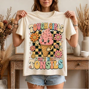 May include: A cream-coloured t-shirt featuring a graphic of a pink ice cream cone with a face, surrounded by colourful flowers and a black and yellow checkerboard pattern. The text reads "TOUGH GIRLS DON'T NEED TONSILS" in a retro, multi-coloured font.