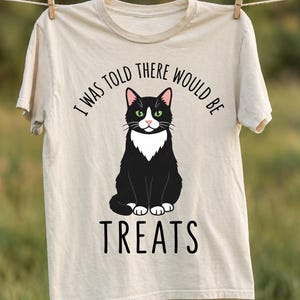Może przedstawiać: Kremowy t-shirt z grafiką czarno-białego kota typu tuxedo siedzącego w pozycji wyprostowanej. Napis "I WAS TOLD THERE WOULD BE TREATS" jest nadrukowany czarnymi wielkimi literami w łuku nad i pod ilustracją kota.