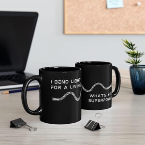 May include: Two black ceramic mugs featuring white text and a graphic of a fiber optic cable. The text on the mugs reads: I BEND LIGHT FOR A LIVING and WHAT'S YOUR SUPERPOWER? The mugs have a smooth finish and a standard handle design.