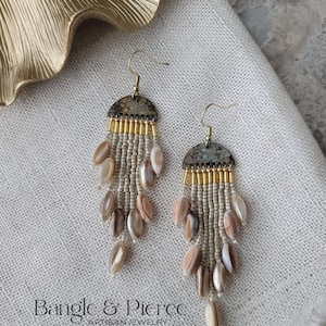 May include: A pair of dangle earrings featuring a semi-circular dark metallic top piece with a textured finish. Below, multiple strands of small cream-colored seed beads are interspersed with polished, oval-shaped iridescent shell beads.