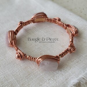 Puede incluir: Un brazalete de alambre de cobre enrollado con varias piedras lisas de color rosa claro de forma irregular y pequeñas cuentas de cobre. El texto Bangle & Pierce Artisan Jewelry está centrado dentro del marco circular del brazalete.