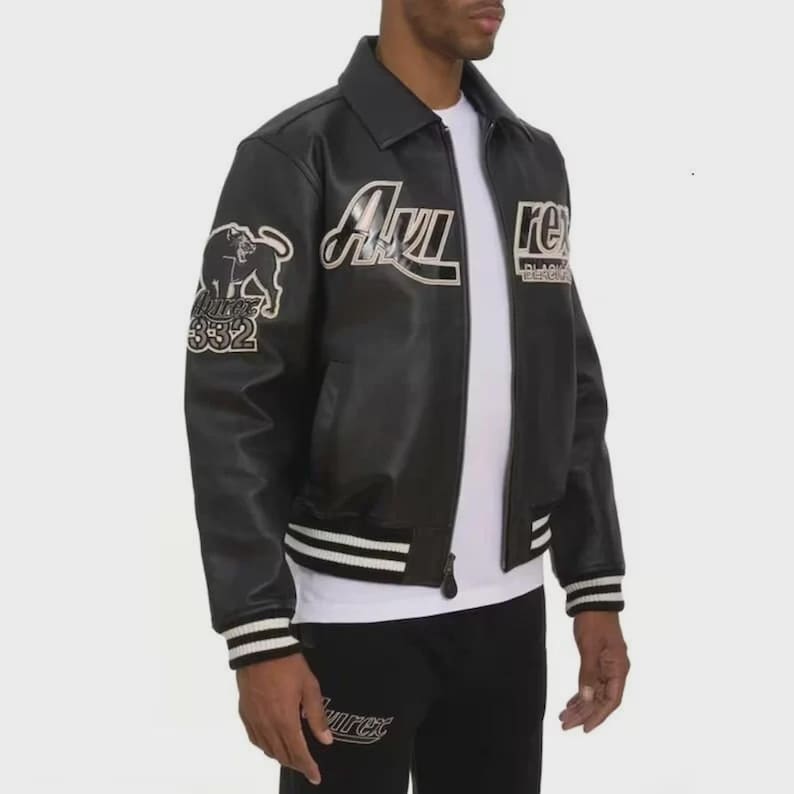 May include: A black leather varsity jacket featuring a white collar, front zipper, and striped ribbed cuffs. The jacket displays white embroidered text reading Avirex and Rex, along with a graphic of a panther and the number 332 on the sleeve.