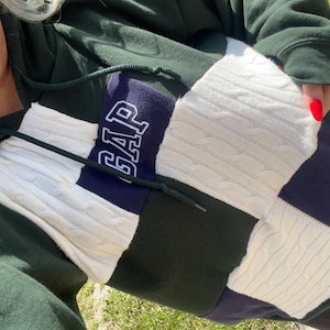 May include: A color-blocked hooded sweatshirt featuring dark green, navy blue, and cream-colored cable knit panels. The center chest displays a navy blue rectangular patch with the word GAP embroidered in white outline lettering and dark green drawstrings.
