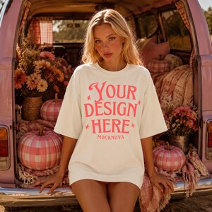 May include: An off-white short-sleeve t-shirt featuring the pink text "YOUR DESIGN HERE" and "MOCKNOVA" printed on the front. The shirt has a crew neckline and a relaxed fit, with small star-shaped accents surrounding the main text.