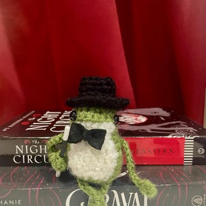 Peut inclure: Une petite figurine de grenouille crochetée verte portant un haut-de-forme noir et un nœud papillon en feutre noir. La grenouille a des yeux en perles noires et tient une baguette magique noire à bout blanc dans une main, posée devant deux livres.