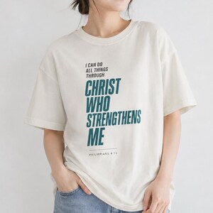 Könnte beinhalten: Ein cremefarbenes T-Shirt mit kurzen Ärmeln und Rundhalsausschnitt, das auf der Vorderseite den schwarzen und blaugrünen Text I CAN DO ALL THINGS THROUGH CHRIST WHO STRENGTHENS ME sowie die Referenz PHILIPPIANS 4:13 in einer serifenlosen Schrift zeigt.