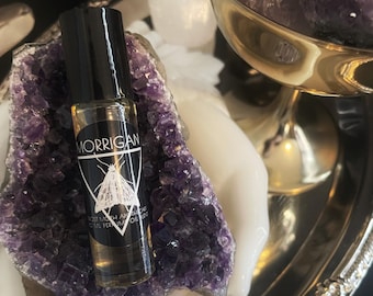 May include: A small glass bottle with a black cap and a white label. The label says "MORRIGAN" and "LOST MOTH AETHERIAL OIL PERFUM OIL BLEND". The bottle is sitting on a purple amethyst crystal.