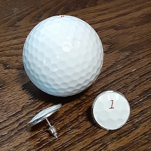 Puede incluir: Una pelota de golf blanca y un par de pendientes con temática de golf. Los pendientes tienen una cara blanca con una textura con hoyuelos y el número "1" en rojo. Los pendientes tienen un poste y un respaldo plateados.