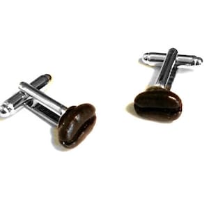 May include: A pair of cufflinks featuring a coffee bean design. The cufflinks have a silver-colored metal bar and a dark brown coffee bean shape. The background is plain white.