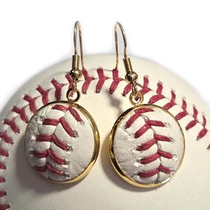 Könnte beinhalten: Baseball-Ohrringe mit weißem und rotem Design und goldfarbenen Haken. Die Ohrringe sind rund und haben ein Baseball-Nahtmuster. Der Hintergrund ist ein Baseball.