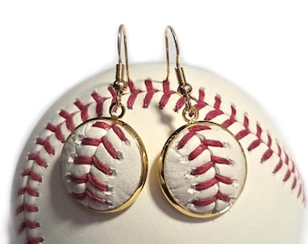 Puede incluir: Pendientes con temática de béisbol con un diseño blanco y rojo, con ganchos dorados. Los pendientes son redondos y tienen un patrón de puntada de béisbol. El fondo es una pelota de béisbol.