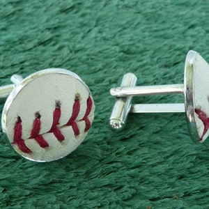 May include: Baseball-themed cufflinks featuring a white leather design with red stitching, mimicking the look of a baseball. The cufflinks have a silver-colored metal border and post, perfect for adding a sporty touch to any outfit.