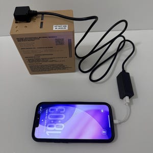 May include: A tan rectangular rechargeable lithium-ion battery with a black power cable connected to a black adapter and a smartphone. The battery features text including "DO NOT USE IN JAVELIN" and technical specifications. The phone screen displays 18:03.