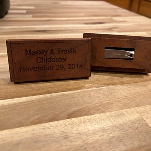 Può includere: Una scatola rettangolare in legno marrone scuro con coperchio scorrevole inciso con il testo Macey & Travis Chidester November 29, 2014. Una chiavetta USB Samsung argentata è parzialmente inserita in una seconda scatola di legno dietro.
