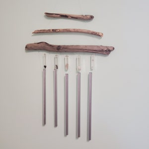 May include: A handcrafted wind chime featuring natural driftwood and clear crystal accents. The chime has five silver-colored tubes suspended below the driftwood, creating a soothing sound. The driftwood pieces are arranged horizontally, adding a rustic touch to the design.