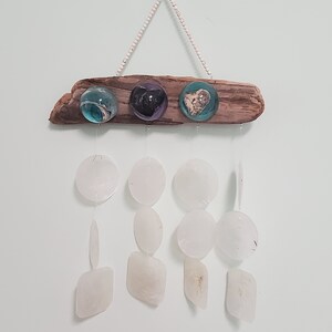 Peut inclure: Un carillon en bois flotté avec trois globes de verre contenant différents objets. Les globes sont suspendus au bois flotté avec des perles blanches. Le carillon a sept cloches en forme de coquille blanche qui pendent.
