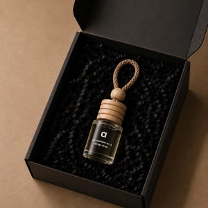 Op de afbeelding: Een klein, doorzichtig glazen flesje met een dop van licht hout en een gevlochten koordlus, liggend in een zwarte doos gevuld met zwart papier. Het flesje heeft een zwart etiket met de witte tekst "a atmosphere no. 6 terrier drive".
