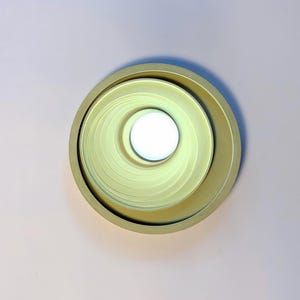 May include: A circular light fixture with a matte pale yellow finish, featuring a recessed design with concentric rings that lead to a bright white circular center light source, creating a layered appearance within the round frame.