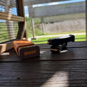 May include: An orange plastic ammunition box with the text 9MM printed on the front, containing brass-cased cartridges. Next to it are two black metal firearm magazines, each loaded with a single brass-cased cartridge visible at the top.