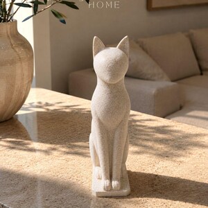 Pode incluir: Uma estatueta de gato em pedra bege salpicada, em posição sentada, com orelhas pontiagudas e um rosto liso sem traços. A escultura tem uma superfície texturizada e assenta numa base retangular. O texto JESSY HOME é visível acima do gato.