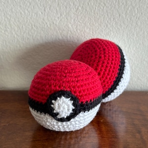 May include: Two spherical crochet toys designed to resemble Poké Balls. Each ball features a red upper hemisphere, a white lower hemisphere, and a black horizontal band encircling the middle with a small white circular center detail.