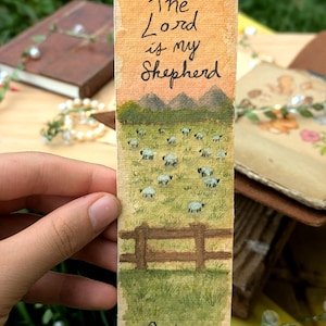 May include: A rectangular bookmark featuring a hand-painted landscape of a green pasture with sheep, a brown wooden fence, and mountains. The text reads The Lord is my Shepherd and Psalm 23 in black ink against an orange and yellow sky background.